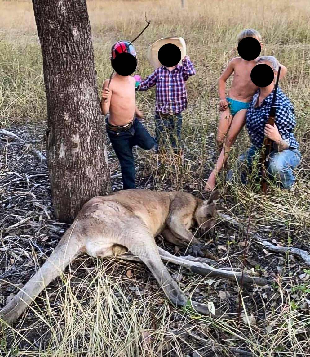 Three young boys pose with with their father holding a gun in front of a dead kangaroo he has killed.