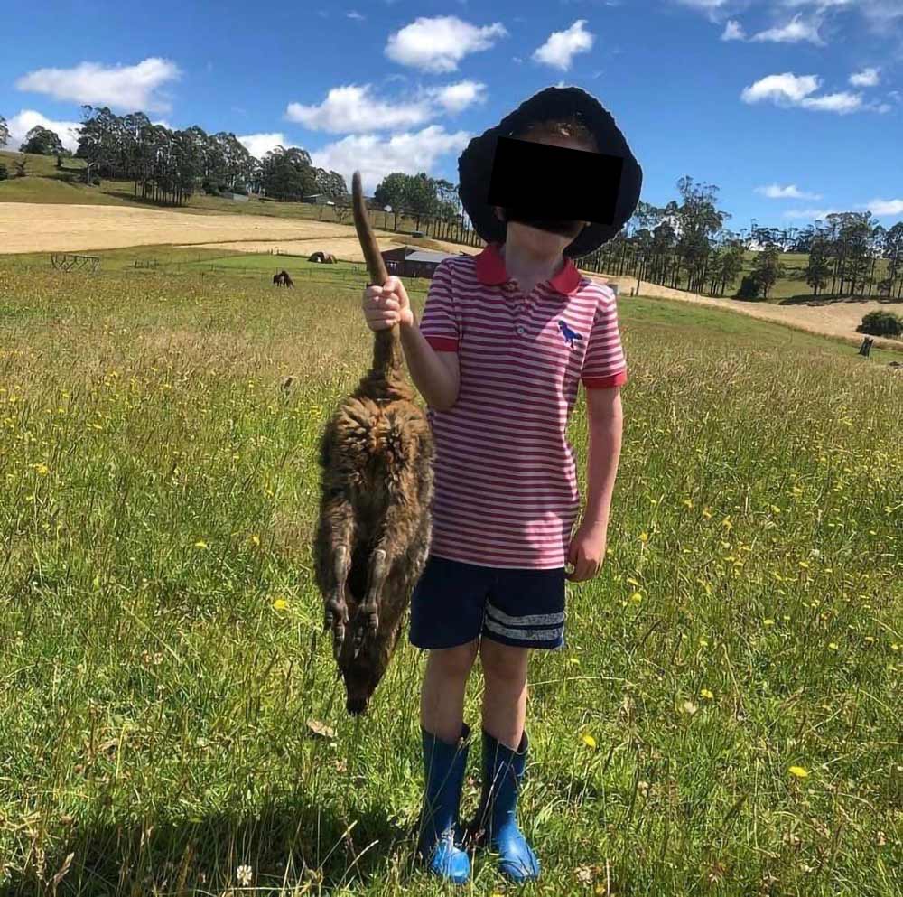 A young boy poses for the camera, holding a dead joey in the air by the tail.